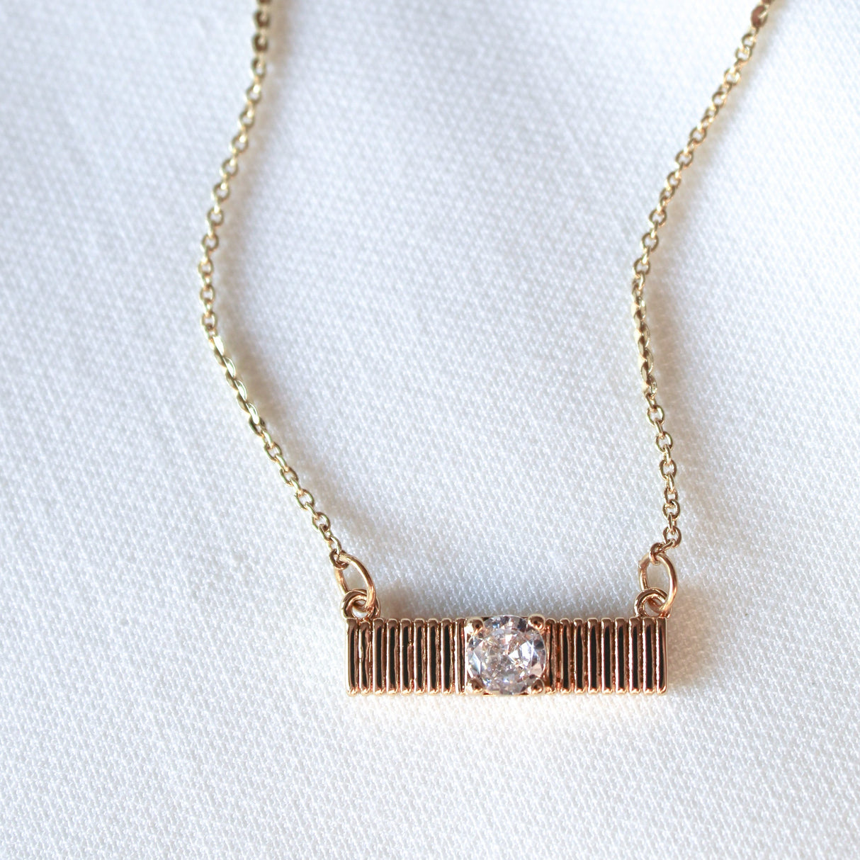 The Quinn Bar Necklace by Kinsey Designs showcases a horizontal gold bar pendant with a central clear gemstone, elegantly displayed on light fabric—perfect as a jewelry gift for any occasion.