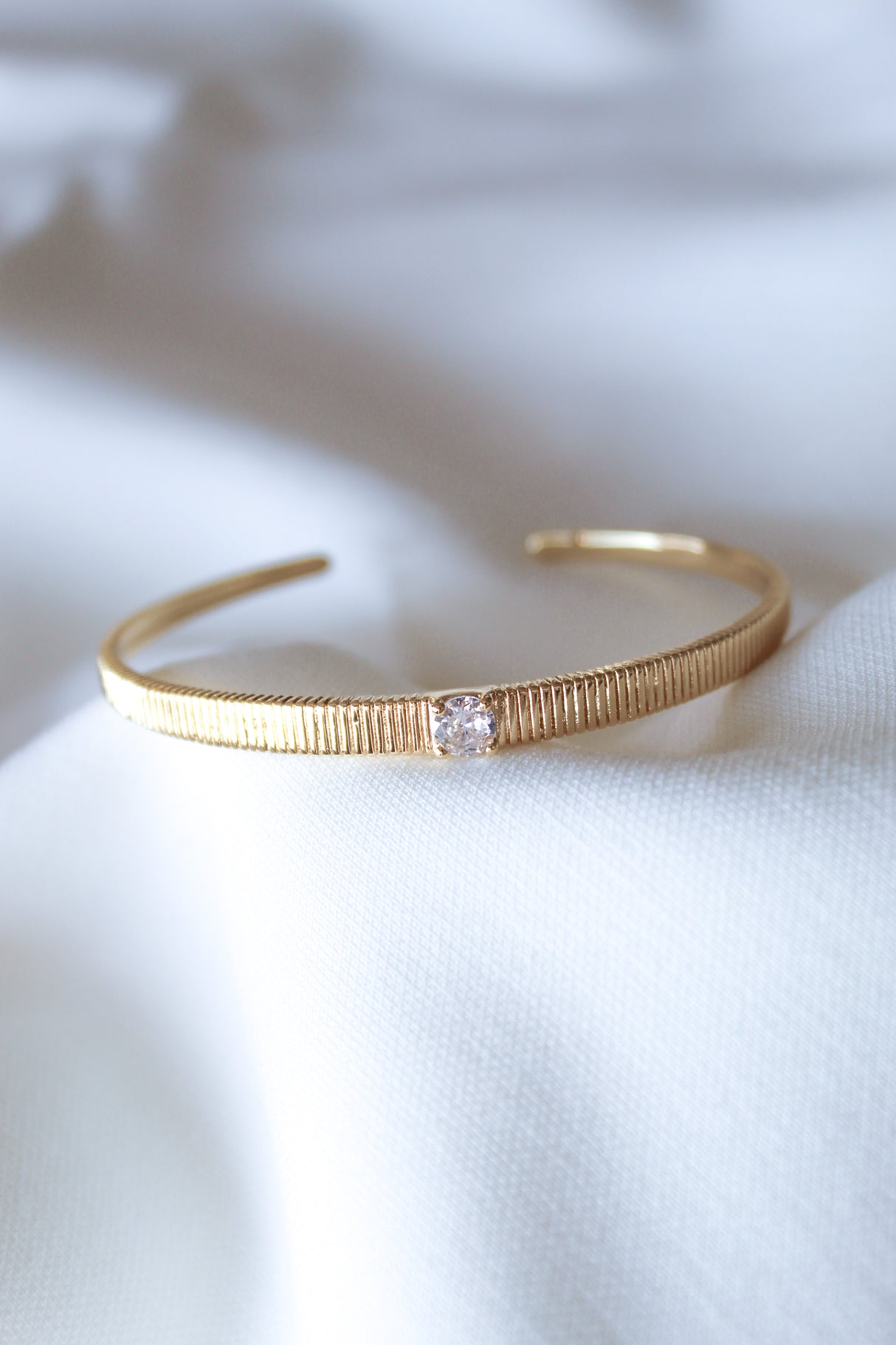 The Quinn Cuff by Kinsey Designs is a gold-filled bracelet featuring a single clear gemstone, shown on soft white fabric.