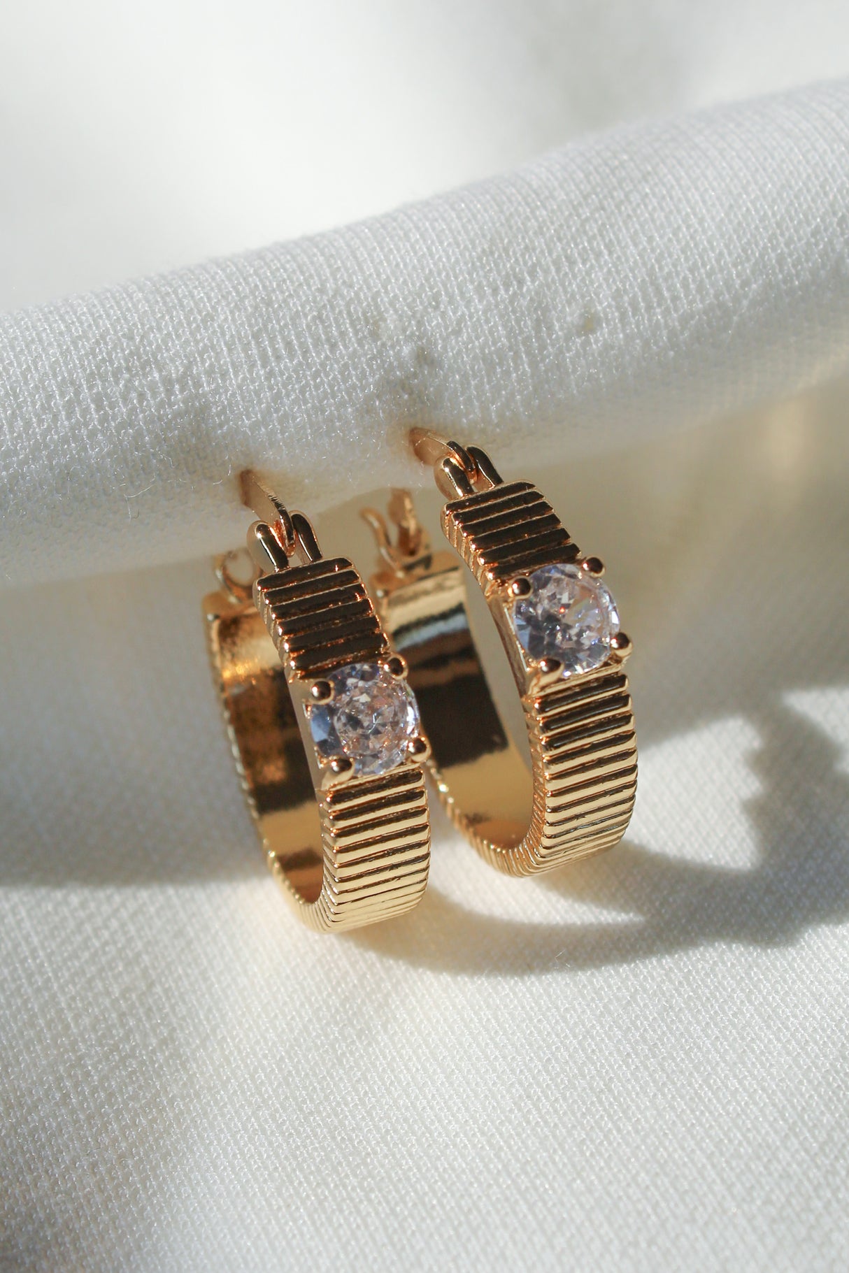 The Quinn Hoop by Kinsey Designs features gold-filled hoops with a ridged design and clear gemstone, .75" in diameter, displayed on white textured fabric.