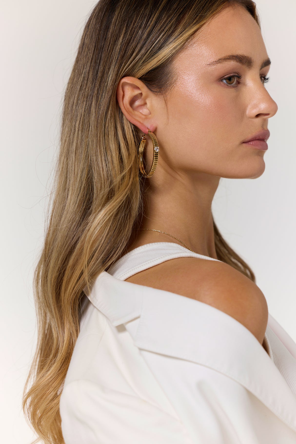 Woman with long blonde hair, wearing a white top and a white jacket off her shoulder, accessorizes with Kinsey Designs Archives’ Quinn Hoop Large gold-filled earrings.