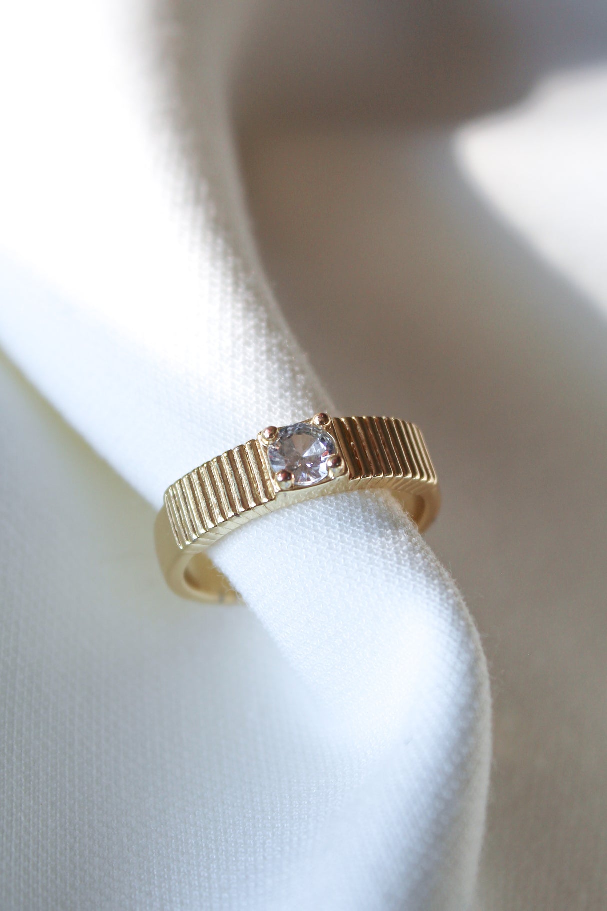 The Quinn Ring by Kinsey Designs features a ridged gold stacking band set with a square-cut CZ stone, elegantly displayed on white fabric.