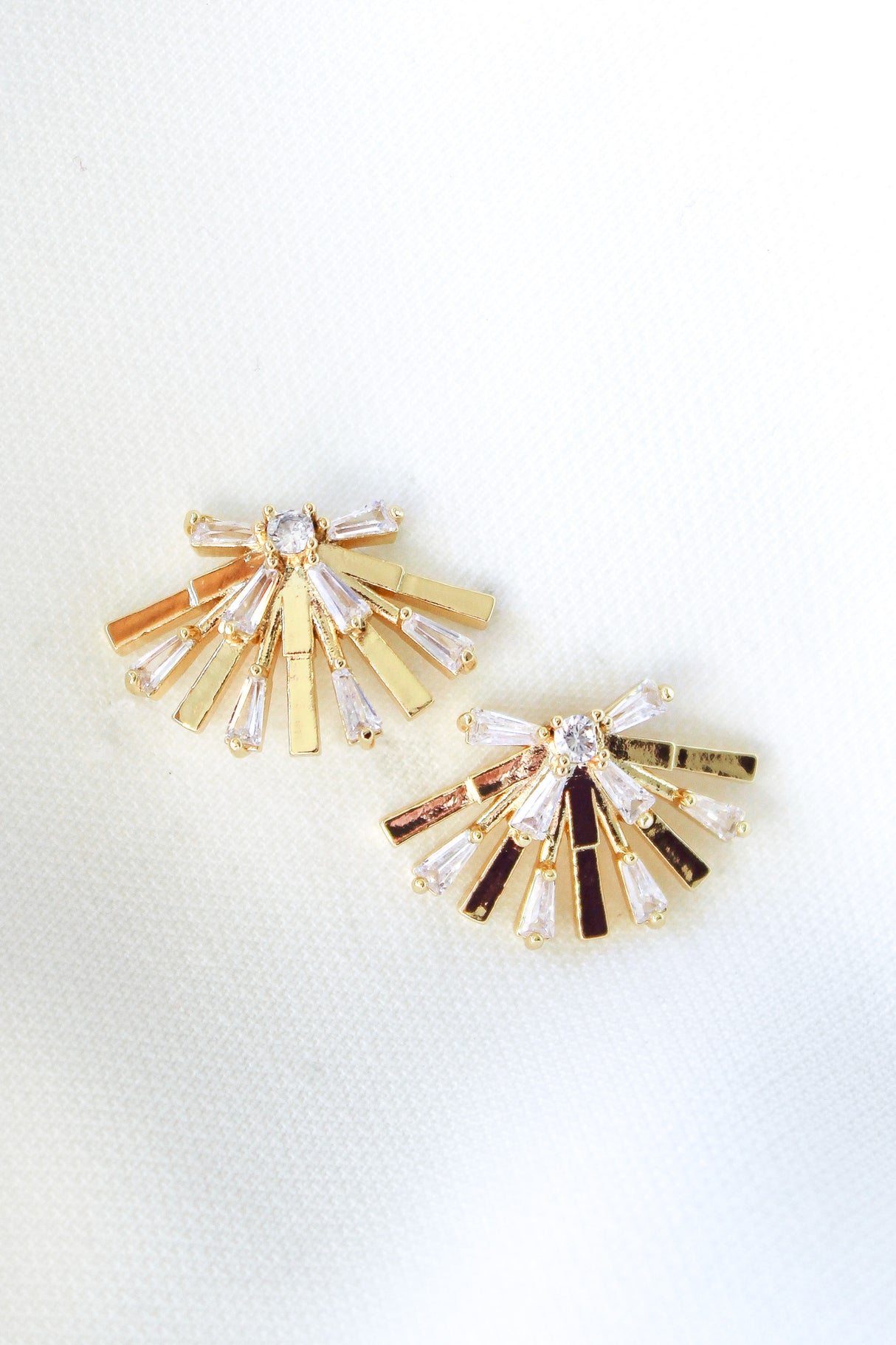 The Rae Post earrings by Kinsey Designs are gold filled, fan-shaped crystal pieces—1 inch long—elegantly displayed on a white textured fabric background, adding sophistication to any jewelry collection.