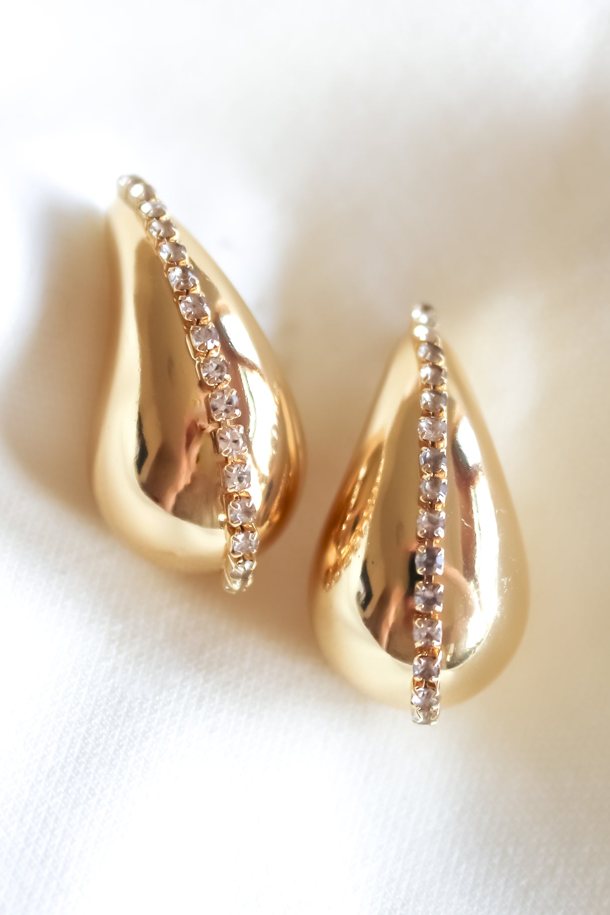 The Kinsey Designs Rhine Teardrop earrings feature gold filled teardrops with a row of crystals, shown on white fabric. Shop these elegant earrings now—final sale.