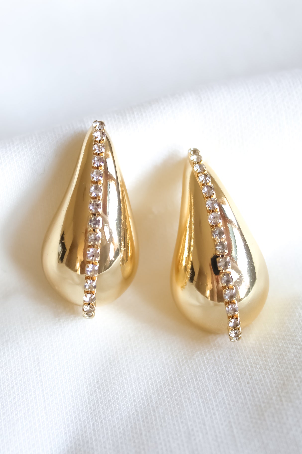 The Rhine Teardrop earrings by Kinsey Designs feature a vertical line of sparkling gems on gold-filled teardrops, elegantly displayed on white fabric. Add refined style to any look—shop them now in our sample sale.