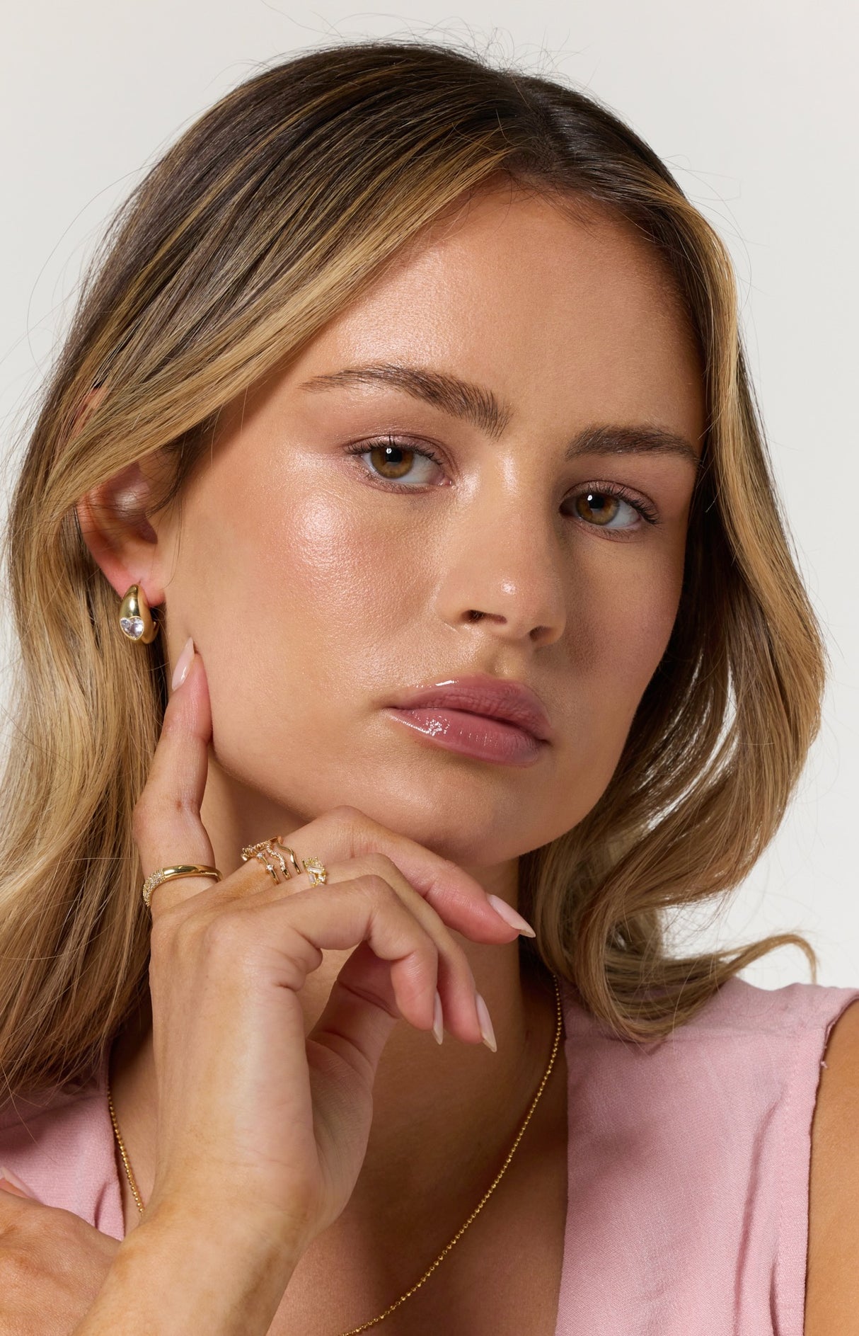 A woman with long blonde hair, wearing the Kinsey Designs Archives Robbie Earring and a pink top, touches her face thoughtfully.