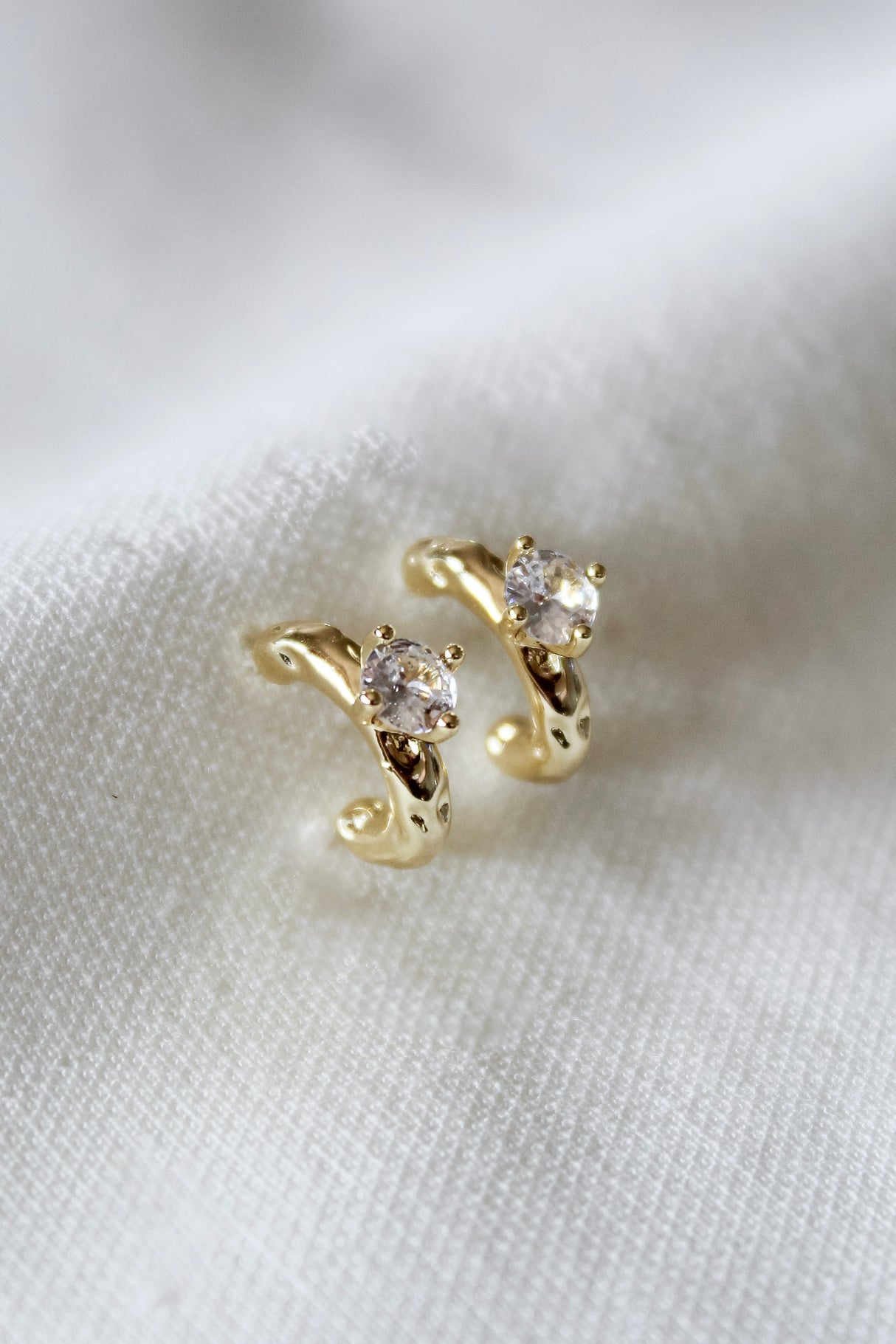 The Kinsey Designs Rugo Hoop earrings, crafted in gold and adorned with clear gemstones, are elegantly displayed on a soft white fabric background.