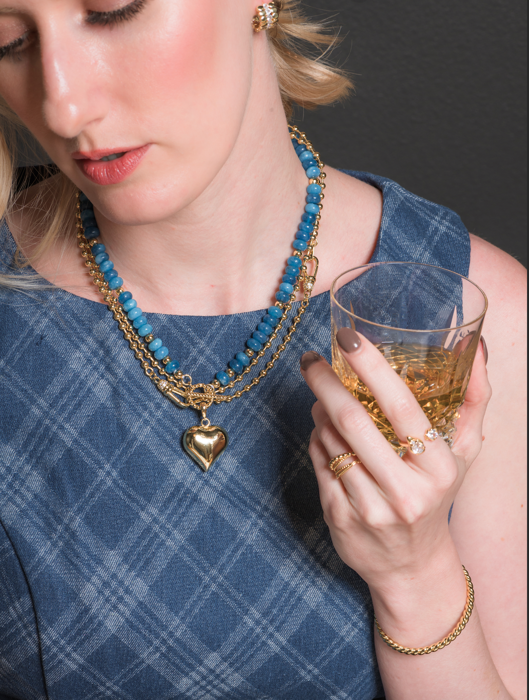 Woman in a blue plaid dress holds a glass of whiskey, wearing gold jewelry and Kinsey Designs’ Aspen Puffy Heart Gemstone Necklace featuring blue sodalite beads and a heart pendant.