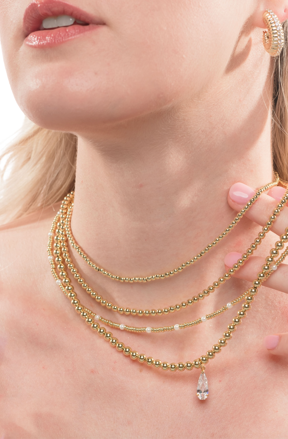 A woman wears layered minimalist jewelry: gold beaded necklaces, including the Kinsey Designs Adda Necklace 4mm with a teardrop pendant, and a gold earring.
