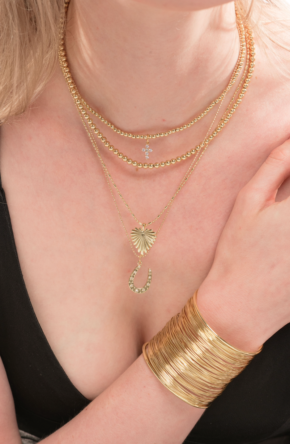 A woman wears layered gold necklaces, featuring a delicate beaded chain and the Kinsey Designs Adda Necklace 5mm, styled with a wide gold bangle and a chic black top for a minimalist jewelry look.
