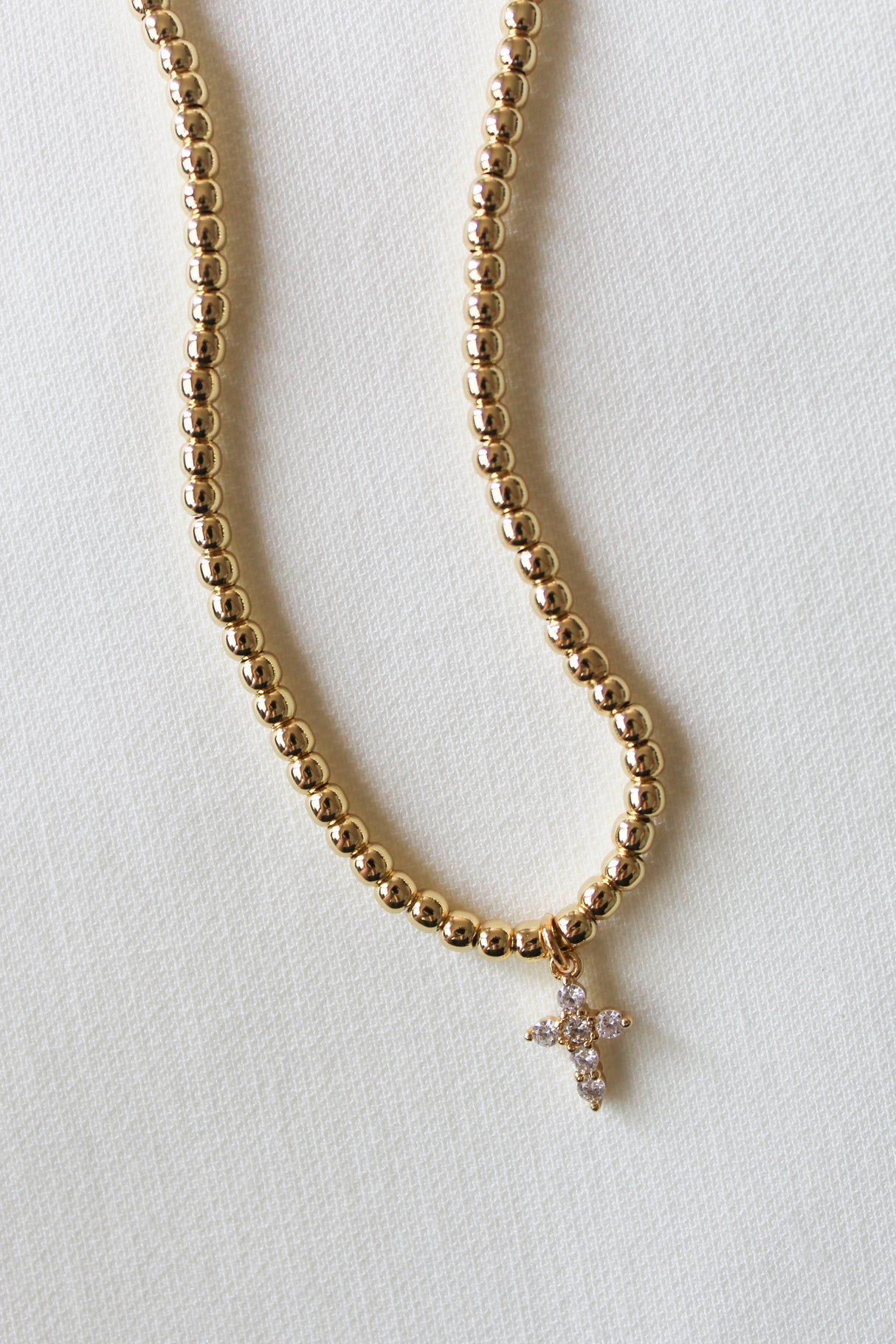 The Selena Necklace by Kinsey Designs features 3mm gold beads and a crystal-studded cross pendant, secured with a clasp and extender, elegantly displayed on a light textured surface.