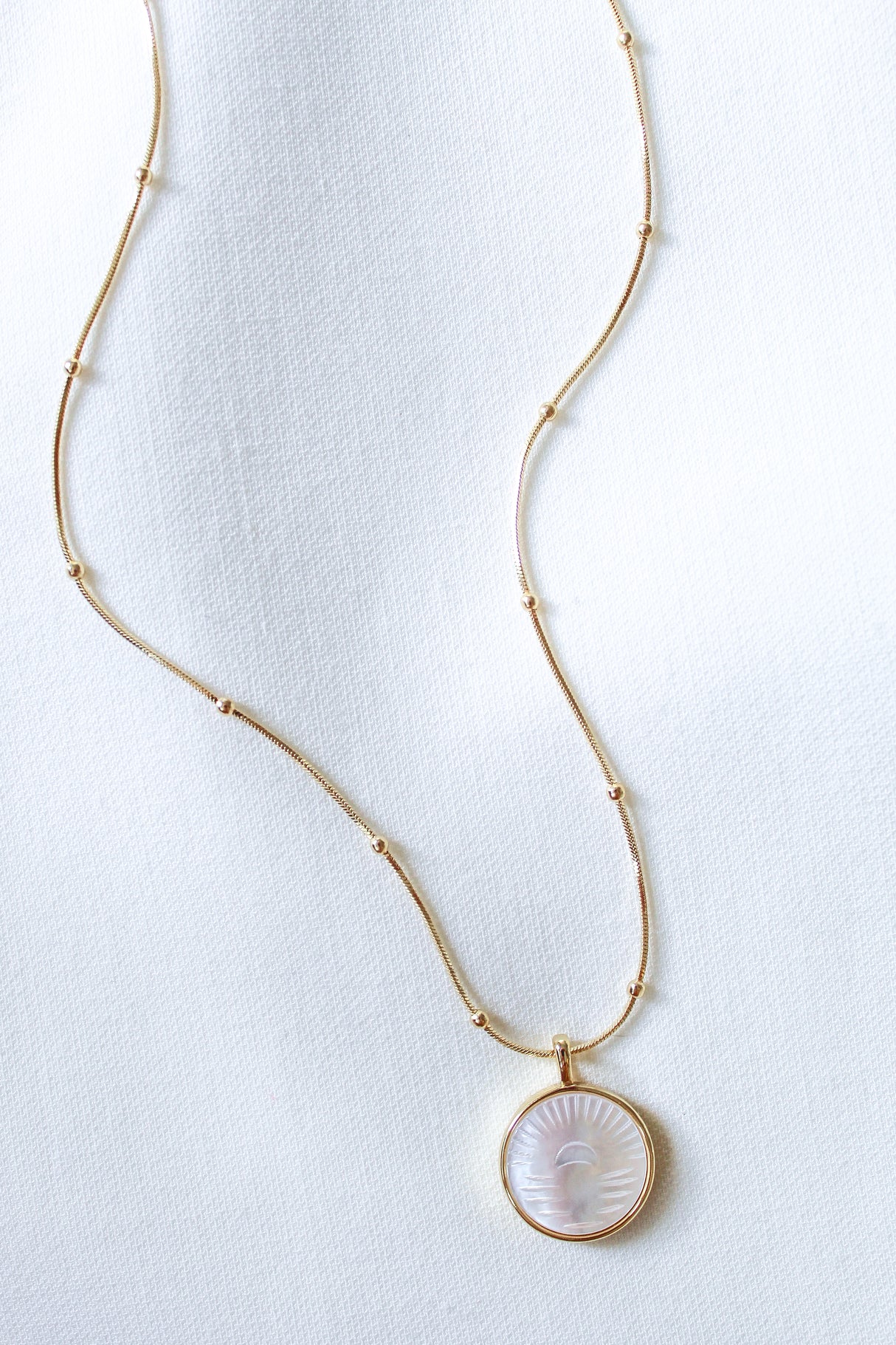 The Shiloh Pearl Necklace by Kinsey Designs Archives features a round, sun-engraved mother-of-pearl pendant on a white textured fabric background.