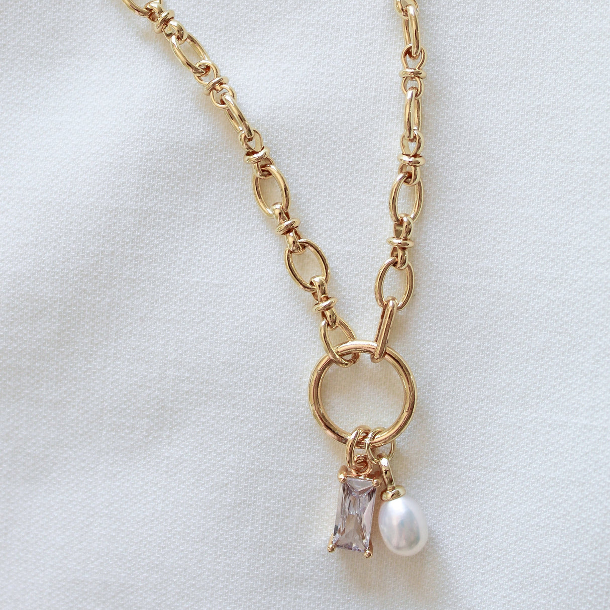 The Sophie Pearl Necklace by Kinsey Designs Archives features a gold-filled chain with a circular pendant, pearl charm, and rectangular cubic zirconia gemstone, elegantly displayed on white fabric.