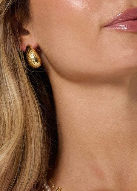 A woman with glossy pink lips and partially visible blonde hair wears Surtur Earrings by Kinsey Designs Archives—shiny, 1-inch-long hammered gold filled earrings.