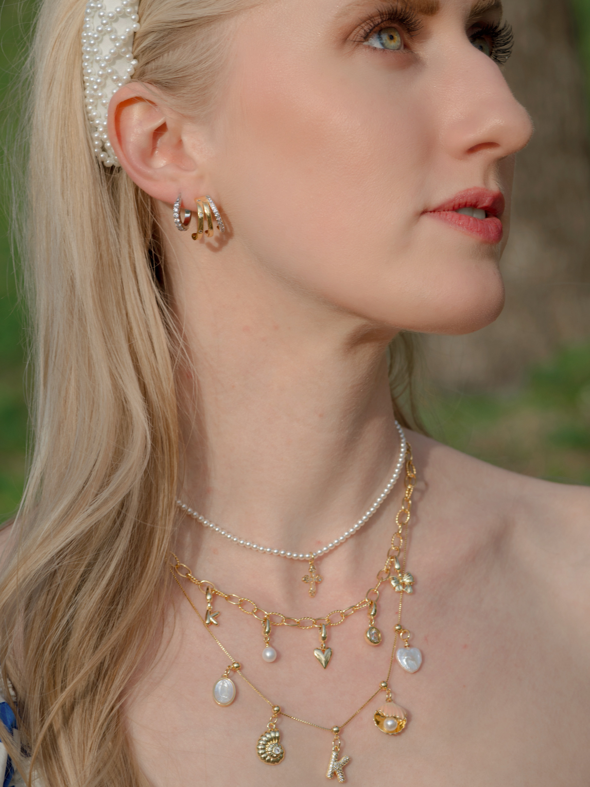 Woman with long blonde hair outdoors wearing layered gold necklaces, including the Kinsey Designs Tilly Cross Necklace, a pearl headband, and gold hoop earrings.