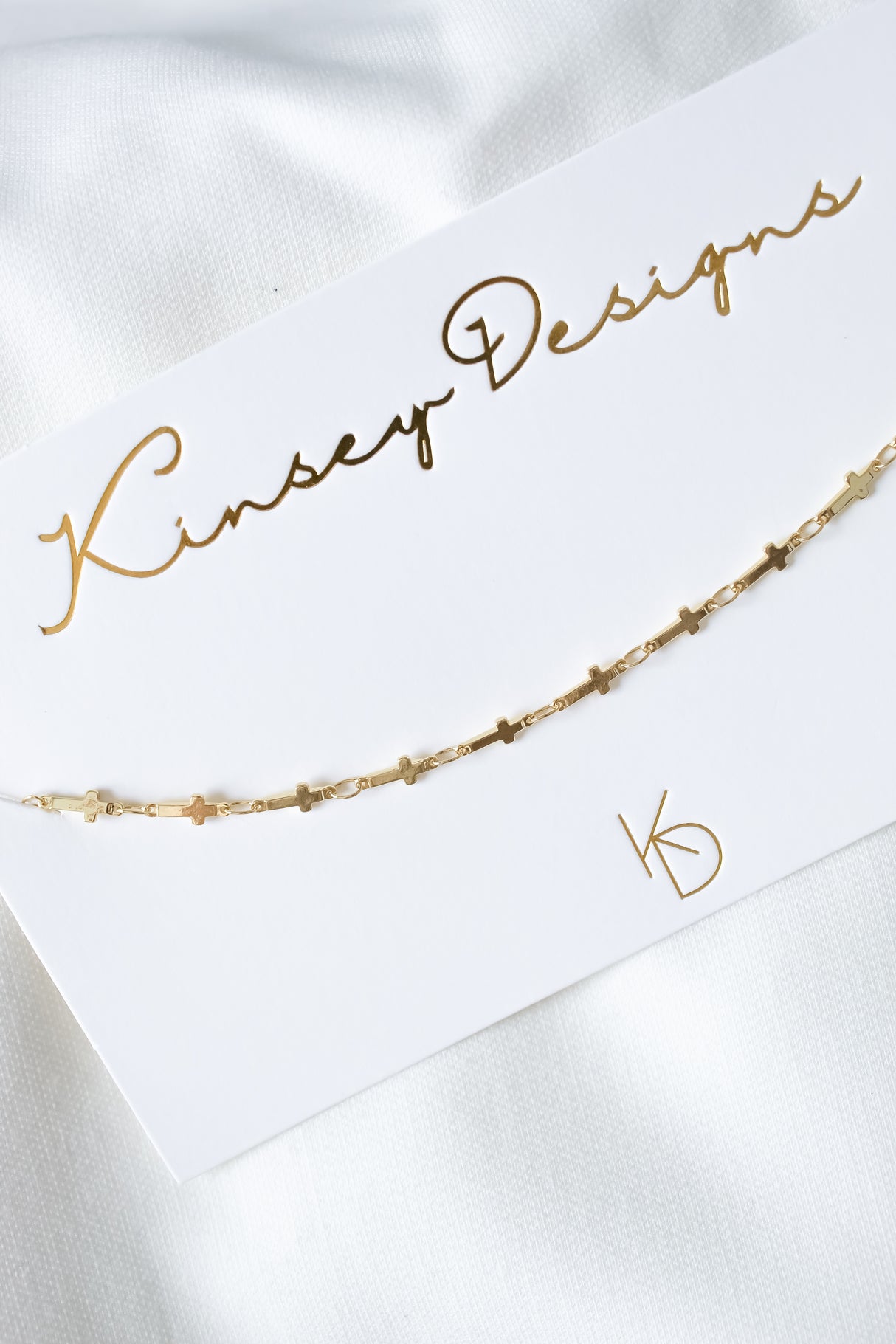 The Trinitee Necklace by Kinsey Designs is displayed on a white card with gold lettering, set atop white fabric.