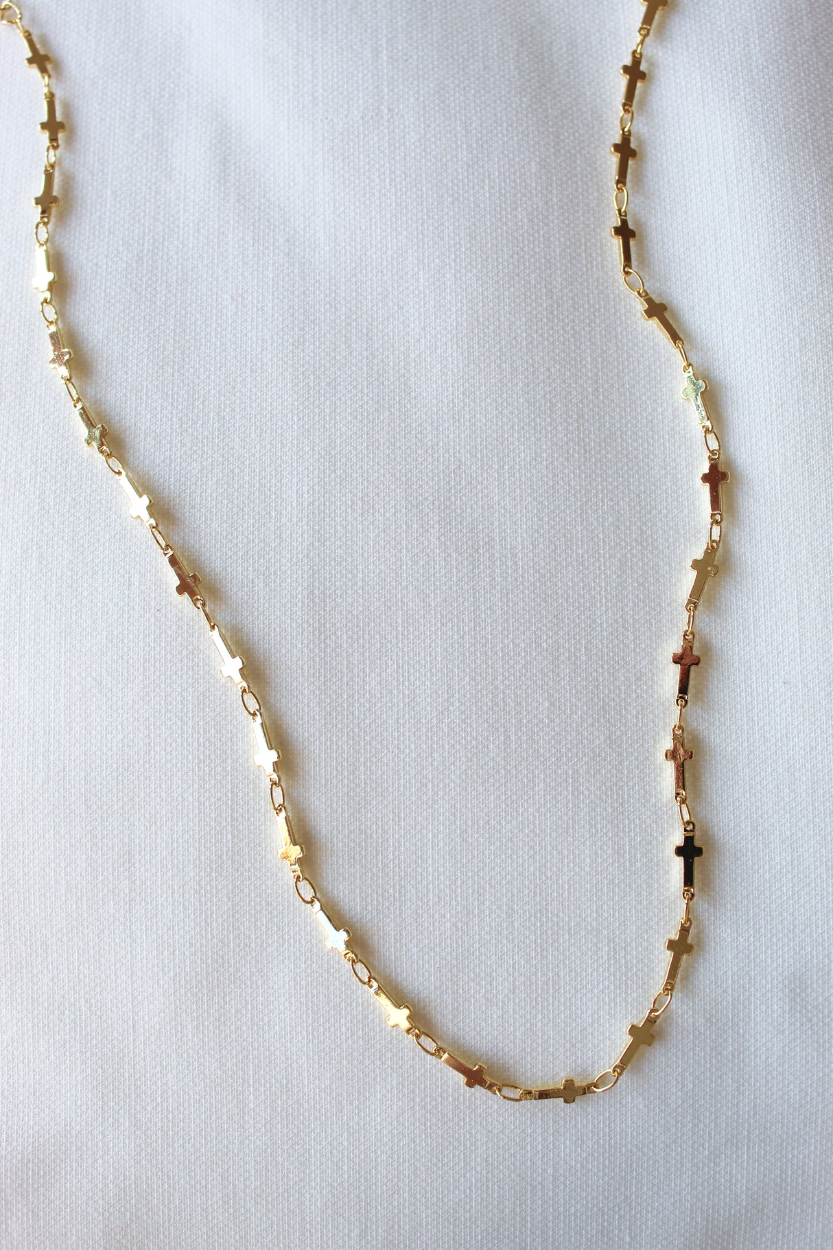 The Trinitee Necklace by Kinsey Designs features small cross-shaped links in gold fill. This elegant 14" piece is ideal for layering or wearing alone, showcased against a white textured background.
