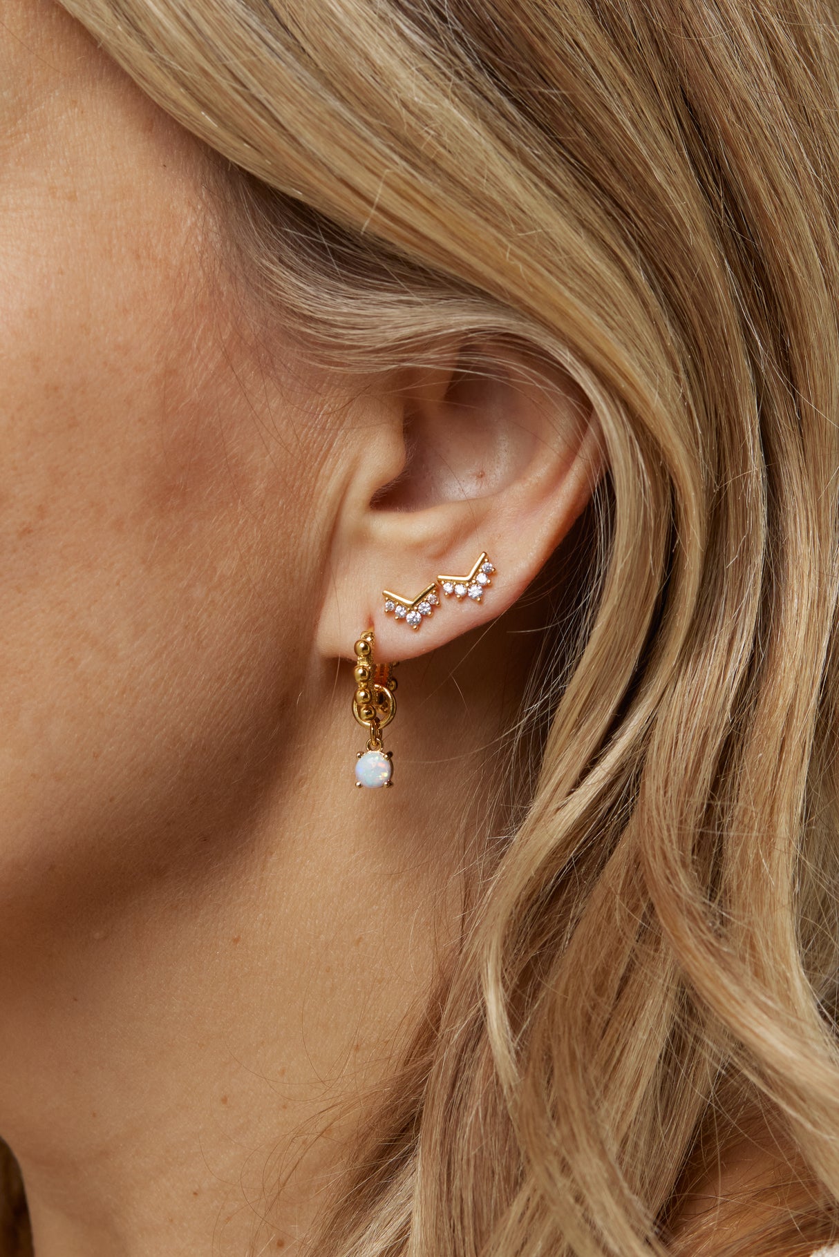 Close-up of a woman's ear with three gold filled earrings from Kinsey Designs Archives, featuring the Villa Stud and a dangling white stone. Add elegance—shop the Sample Sale now!.
