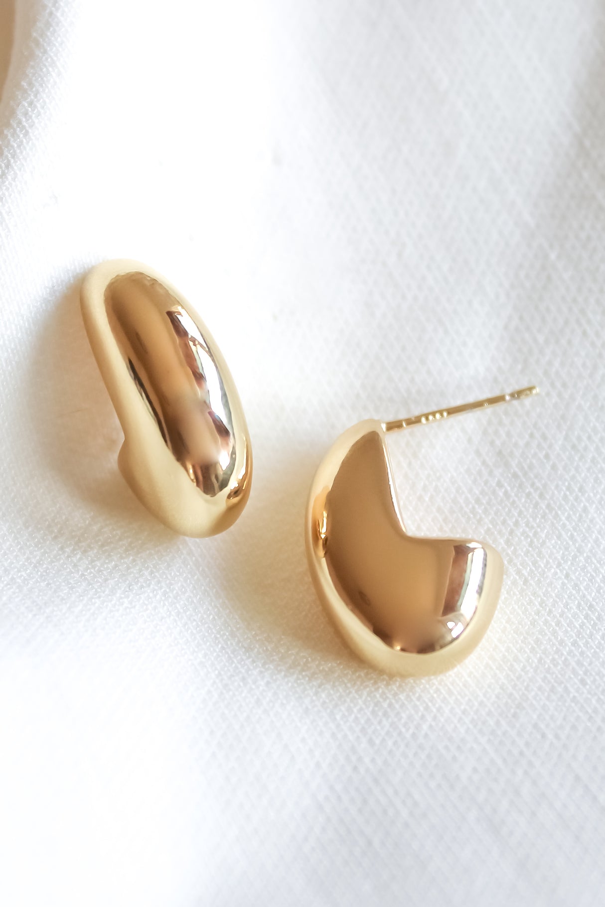 The Weston Earrings by Kinsey Designs—shiny, chunky gold filled pieces—rest on white textured fabric. Elevate your style and don’t miss your chance to grab these at our exclusive sample sale!.
