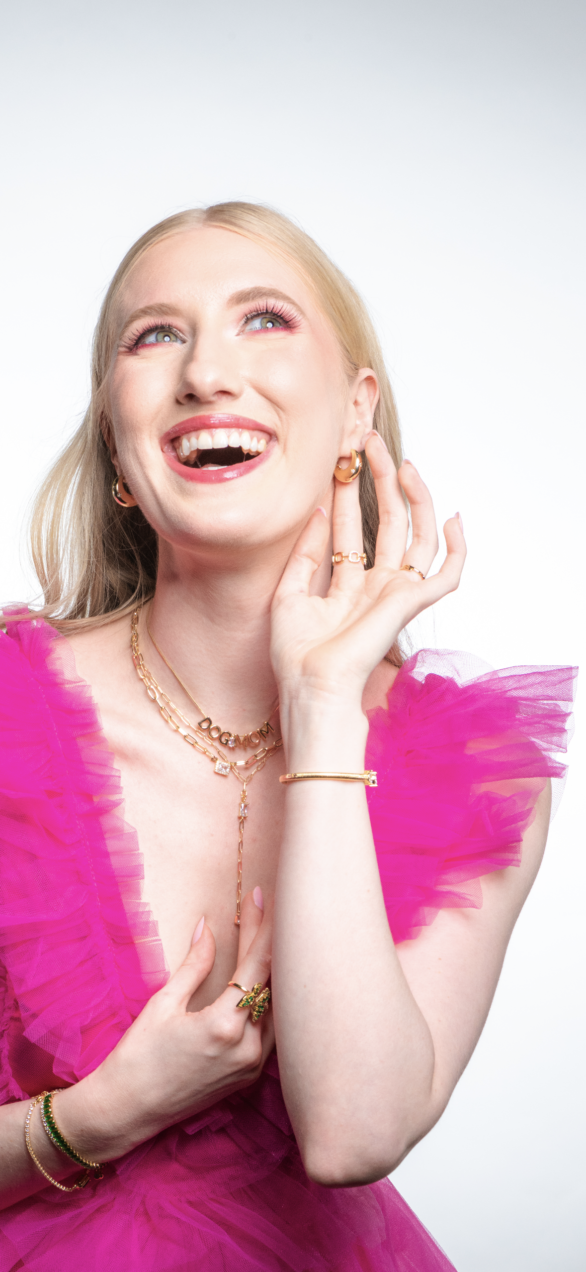 A smiling woman in a bright pink dress wears gold jewelry, featuring the stunning Kinsey Designs Archives Yara Lariat gold-filled cubic zirconia necklace, as she touches her face and looks upward.