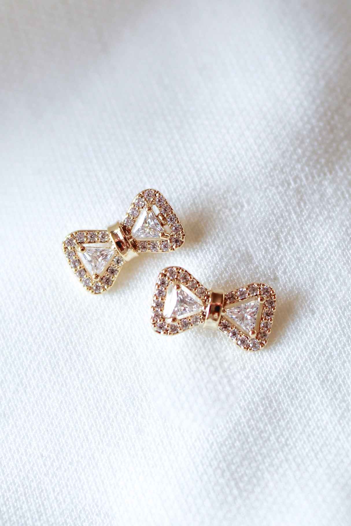 The Kinsey Designs Zoe Bow Stud earrings feature a gold-filled bow shape with small crystals, displayed on a white textured fabric background.