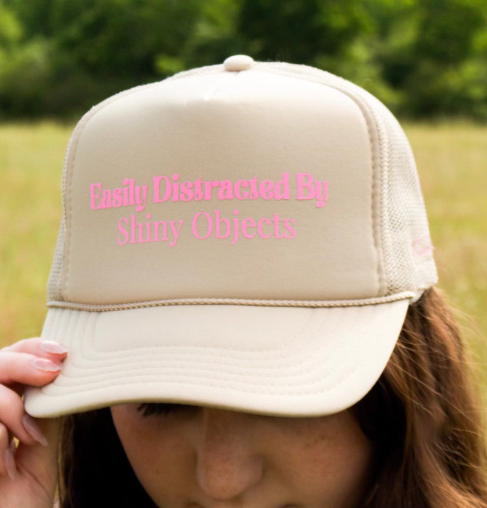 Wearing Kinsey's Shiny Objects Hat by Kinsey Designs Archives, featuring pink "Easily Distracted By Shiny Objects" text, this adjustable beige cap is the perfect summer accessory for lakeside adventures.