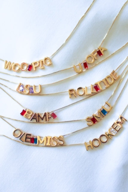 Show your college spirit with Kinsey Designs Gold Letter Slide Charms necklaces, featuring colorful gems on a white cloth background. Personalize with initials or create a custom necklace for a unique look.