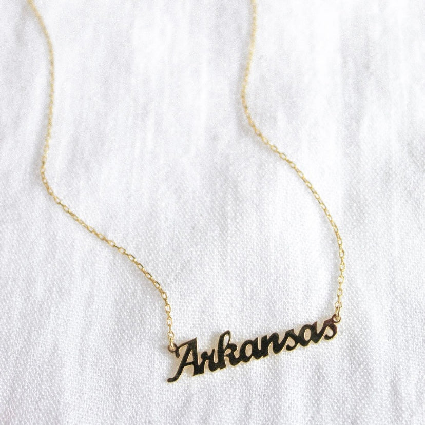 The State of Mind Necklace by Kinsey Designs Archives features a gold filled "Arkansas" cursive pendant, shown on a white fabric background.