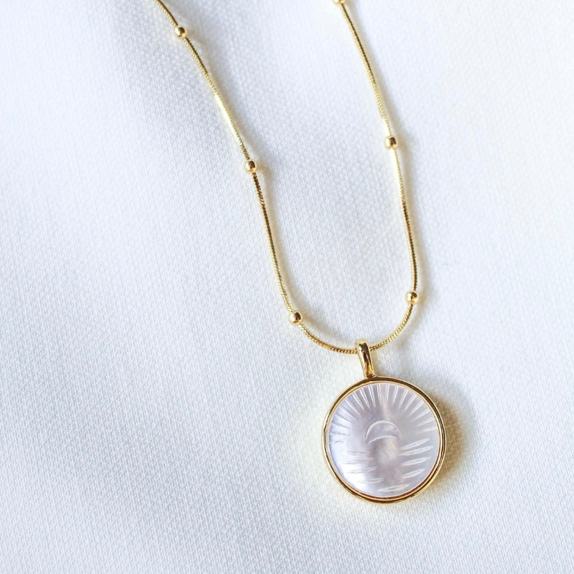 The Shiloh Pearl Necklace by Kinsey Designs Archives showcases a gold filled chain with a round, carved mother-of-pearl face pendant, elegantly displayed on white fabric.