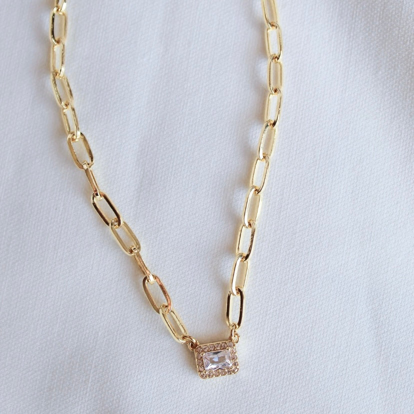 The Posh Necklace by Kinsey Designs features gold filled rectangular links and a square crystal pendant, elegantly showcased on white fabric—an ideal statement piece for any occasion.