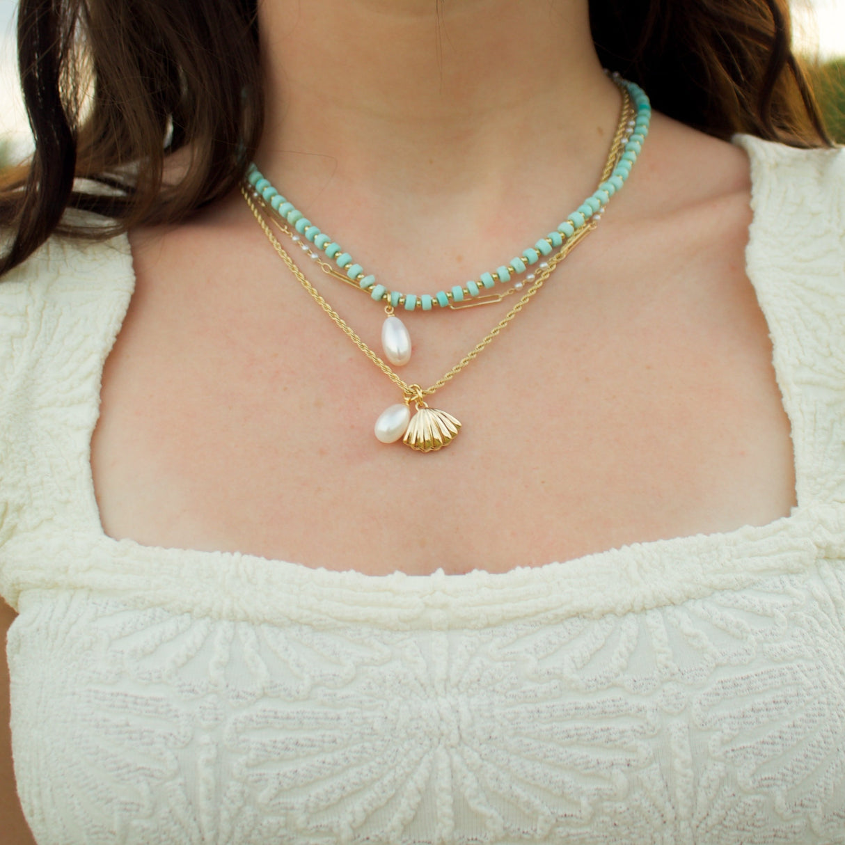 A woman wears a white textured top styled with the Cove Pearl Necklace by Kinsey Designs Archives, layered with turquoise and gold chain necklaces.