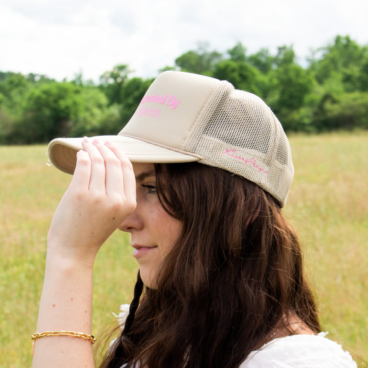In a grassy field, a woman wearing Kinsey's Shiny Objects Hat by Kinsey Designs Archives adjusts the beige mesh cap and gazes into the distance—an ideal summer accessory with adjustable sizing.