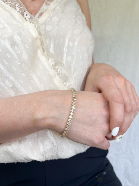 A person wears a lacy white top, black pants, manicured white nails, and a Kinsey Designs Archives Gator Bracelet—subtly styled with its gold finish and adjustable 6.5-inch chain plus extender for the perfect fit.