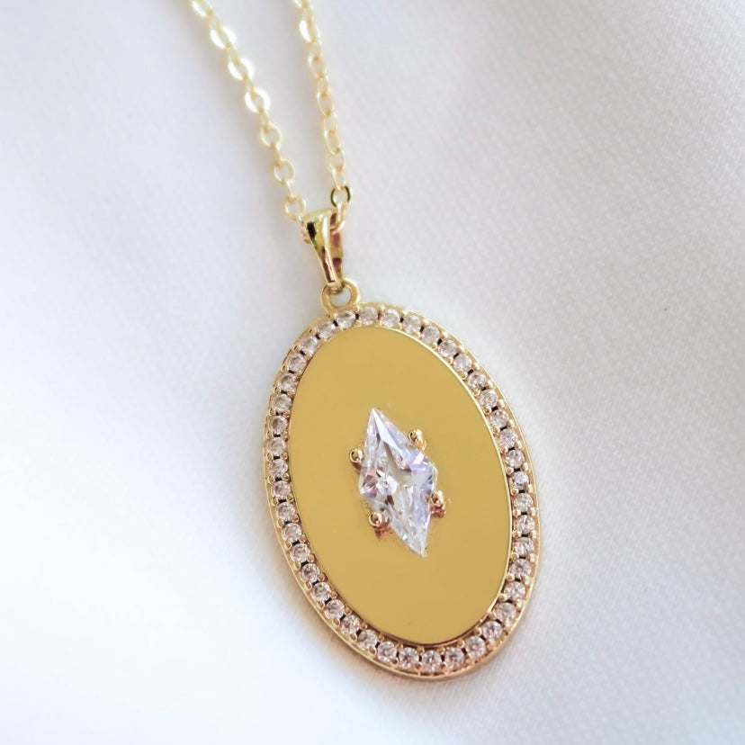 The Fique Necklace by Kinsey Designs features a gold-plated oval cubic zirconia pendant with a marquise-cut center and diamond accents on the edge. Displayed on white fabric, its 32-inch chain adds elegance to this stunning piece.