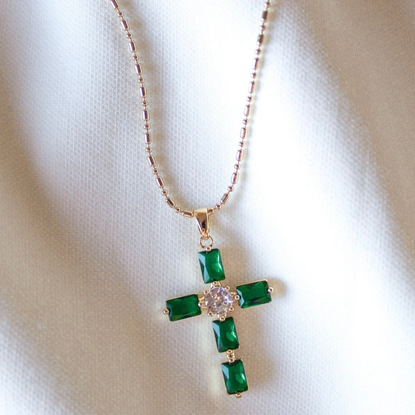 The Tepri Necklace by Kinsey Designs Archives features a gold filled cross pendant with green stones and a central clear gem, displayed on white fabric. Includes a 19-inch chain—perfect for any gold filled jewelry collection.