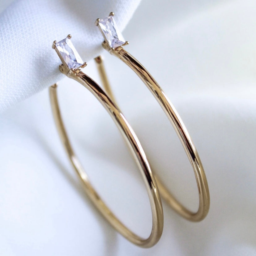 The Darra Hoop Large by Kinsey Designs Archives features gold-plated hoops with rectangular cubic zirconia stones, shown on a white fabric background.