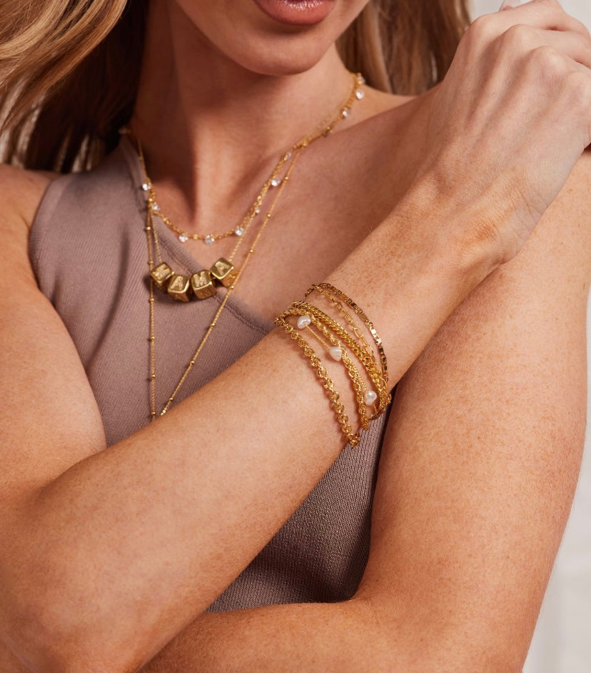 A woman in a sleeveless taupe top showcases layered gold plated chain necklaces and the Emmy Bracelet by Kinsey Designs Archives.