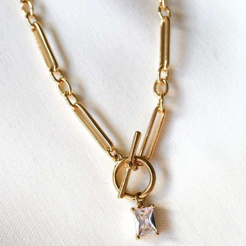 The Gilmore Necklace by Kinsey Designs is a gold-filled toggle pendant necklace with a square-cut clear gemstone, showcased on a white fabric background.