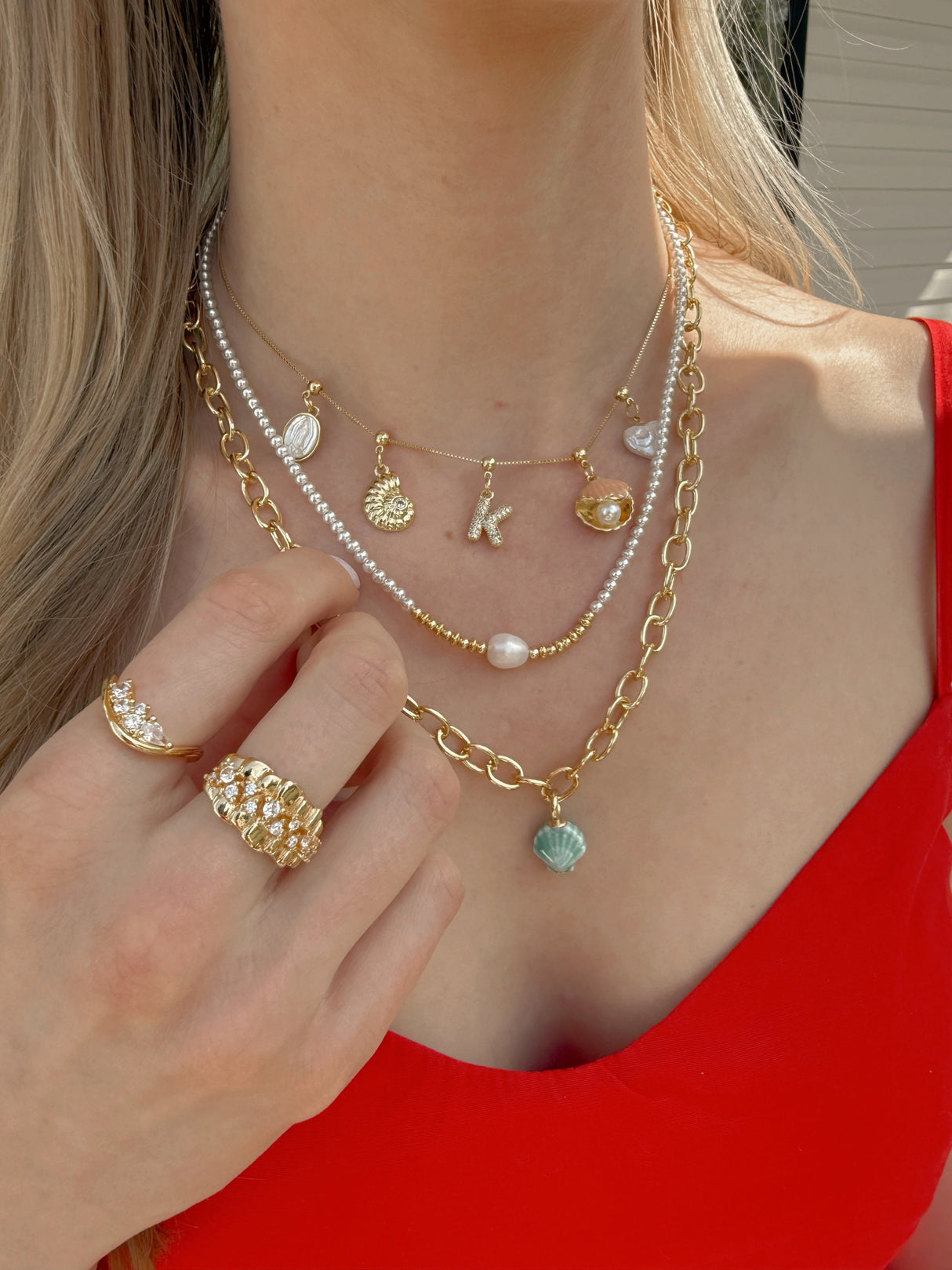 A woman in a red top wears layered gold necklaces, Kinsey Designs' Charm Bar Necklaces, rings, and the Virgin Mary Grip Charm, with her hand touching her chest.