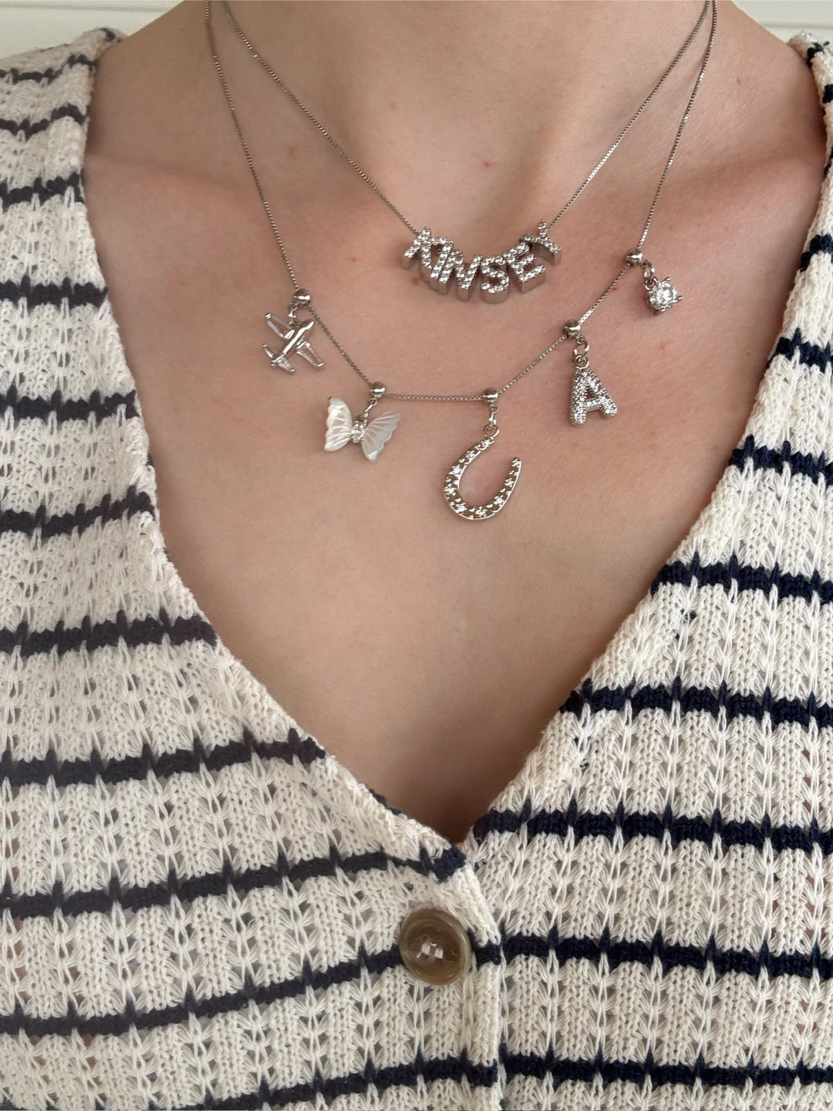 Close-up of three layered Kinsey Designs Charm Bar Necklaces featuring Silver Birthstone Grip Charms, styled over a cream knit top with black stripes and a button.
