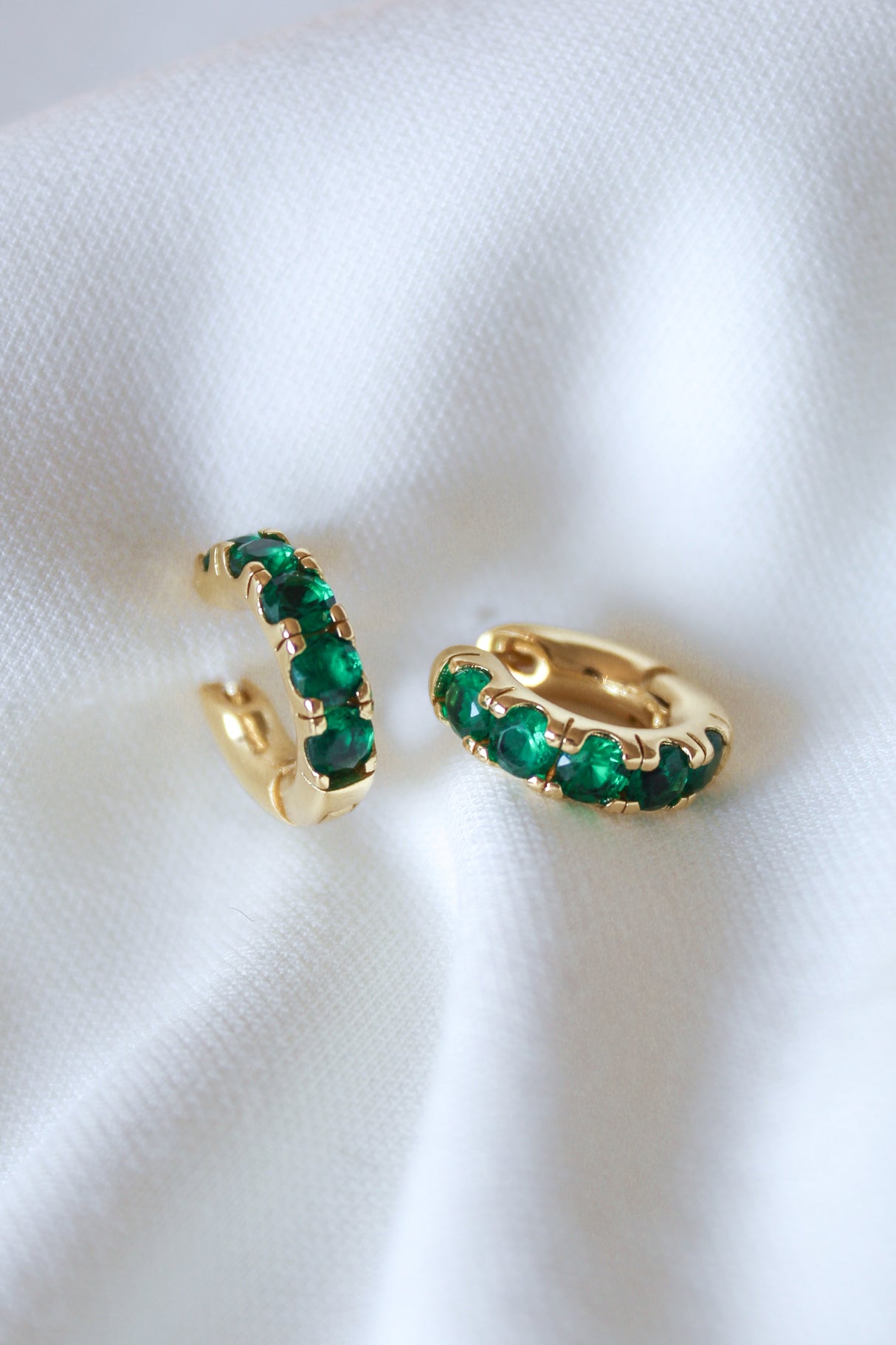 The Abra Huggie by Kinsey Designs Archives features gold-filled hoops with green gemstones (.65" diameter) displayed on soft white fabric—an elegant, eye-catching jewelry piece.