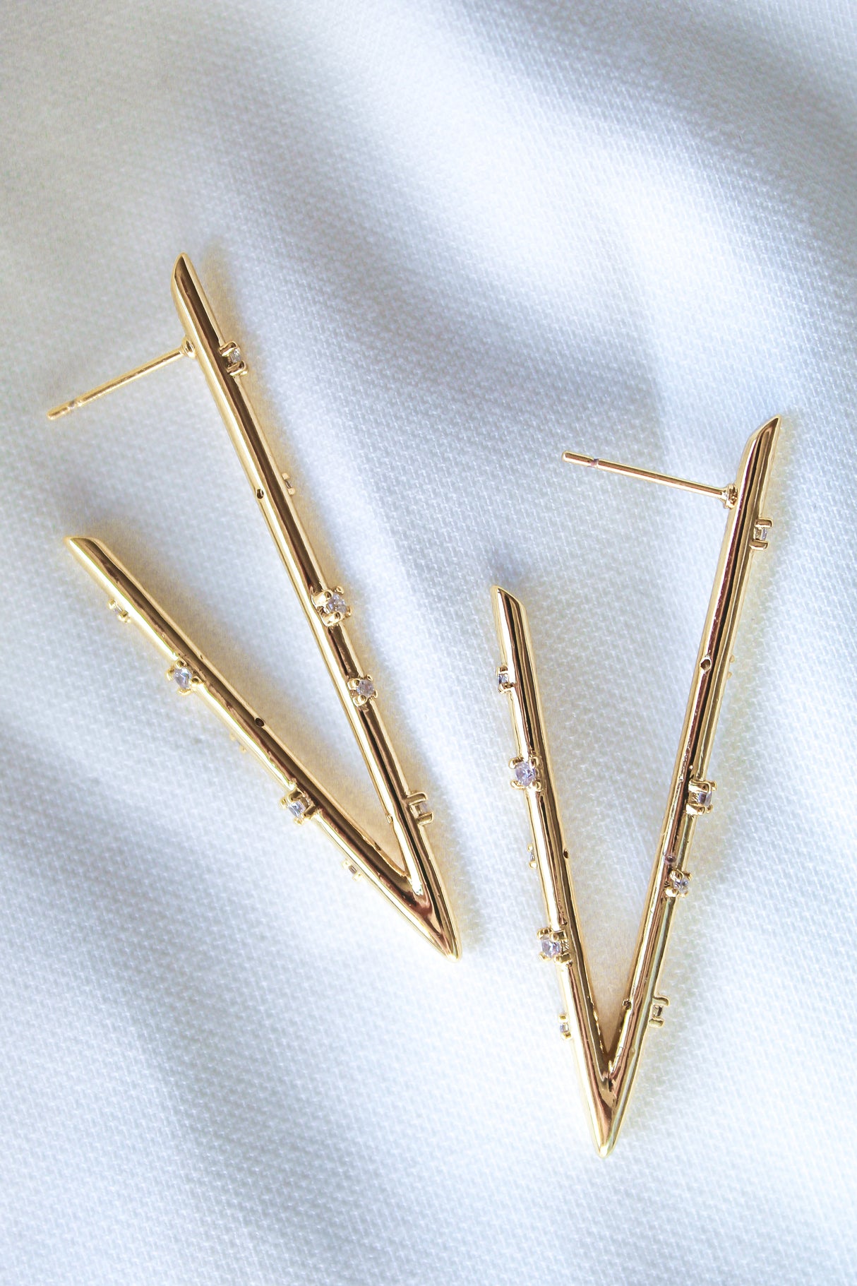 The Bowen Earring Luxe by Kinsey Designs features gold-filled V-shaped earrings with small cubic zirconia gemstones, shown on a white textured fabric background.