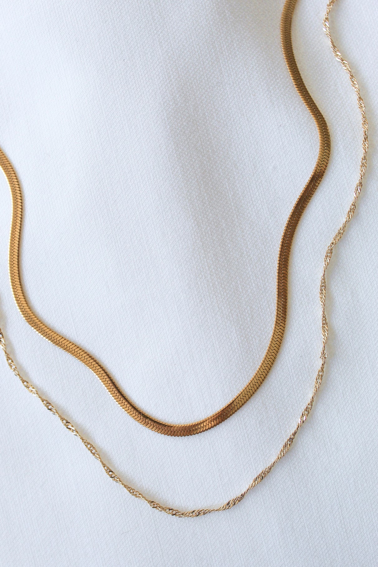 The Callum Layer by Kinsey Designs Archives features two 14k gold-filled necklaces—one flat, one twisted—styled as a layered necklace on white fabric. Both include a 3" extender for adjustable length.
