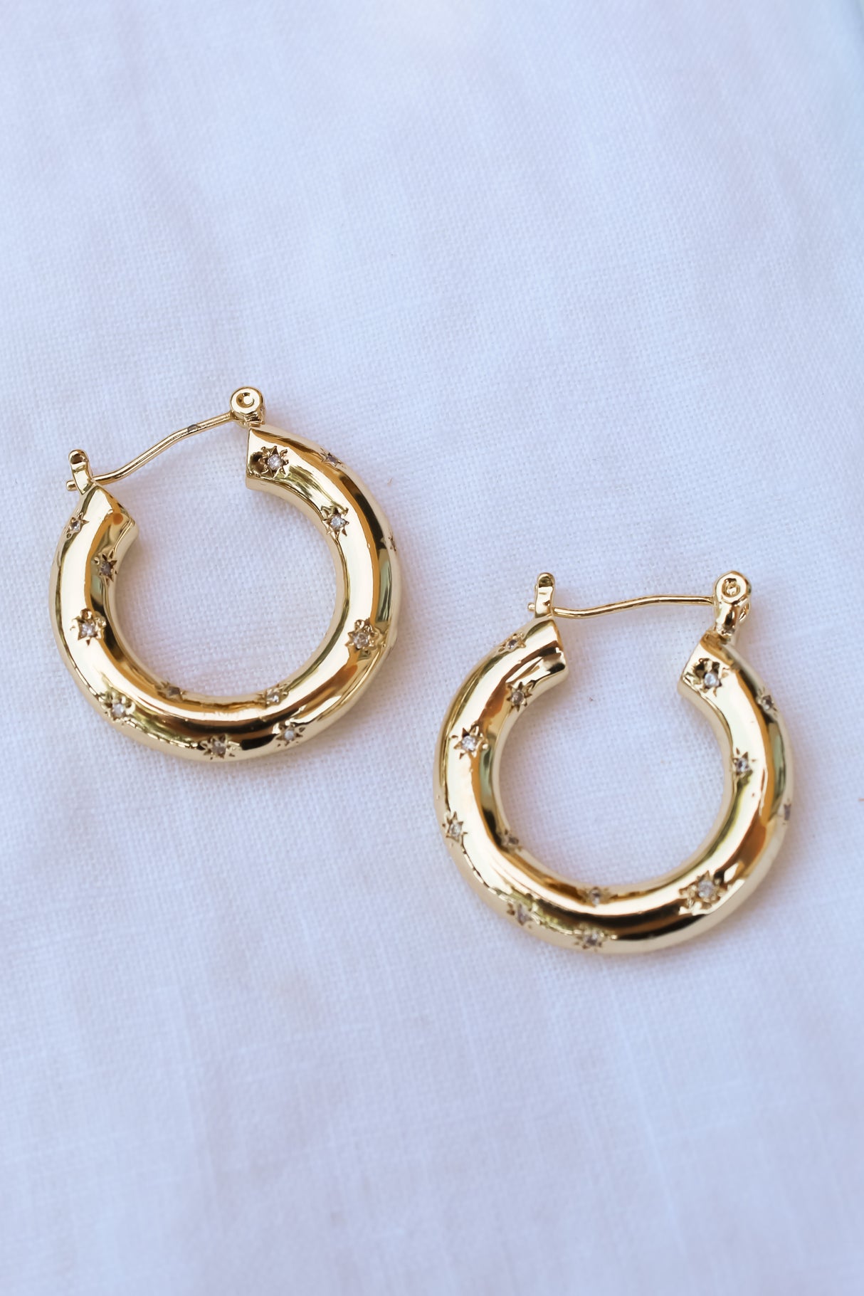 The Camber Earring by Kinsey Designs Archives is a 14k gold filled hoop with small engraved stars and a 1.25-inch circumference, shown on a white fabric background.