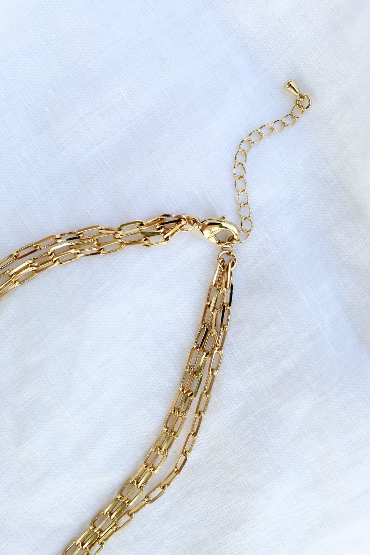The Carmen Triple Layer necklace by Kinsey Designs Archives features three elegant gold-filled strands and an adjustable clasp, designed as a 14-inch piece with extender, displayed on a white fabric background.