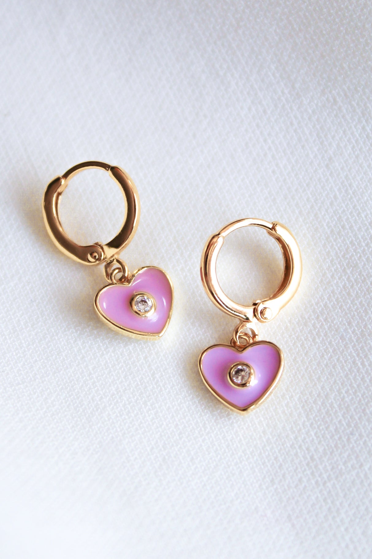 The Cutie Huggie by Kinsey Designs Archives features 1-inch gold filled earrings with pink heart-shaped charms and a central gem, shown on a light fabric background.