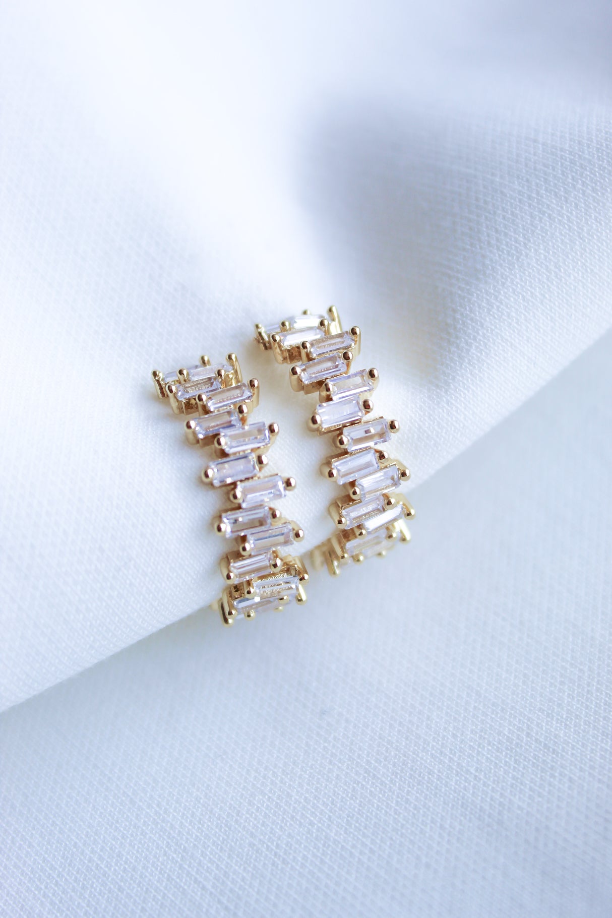 The Ettie Hoop by Kinsey Designs Archives are gold-filled earrings with a 1" diameter, featuring rectangular cubic zirconia stones in a zigzag pattern, shown on white fabric.