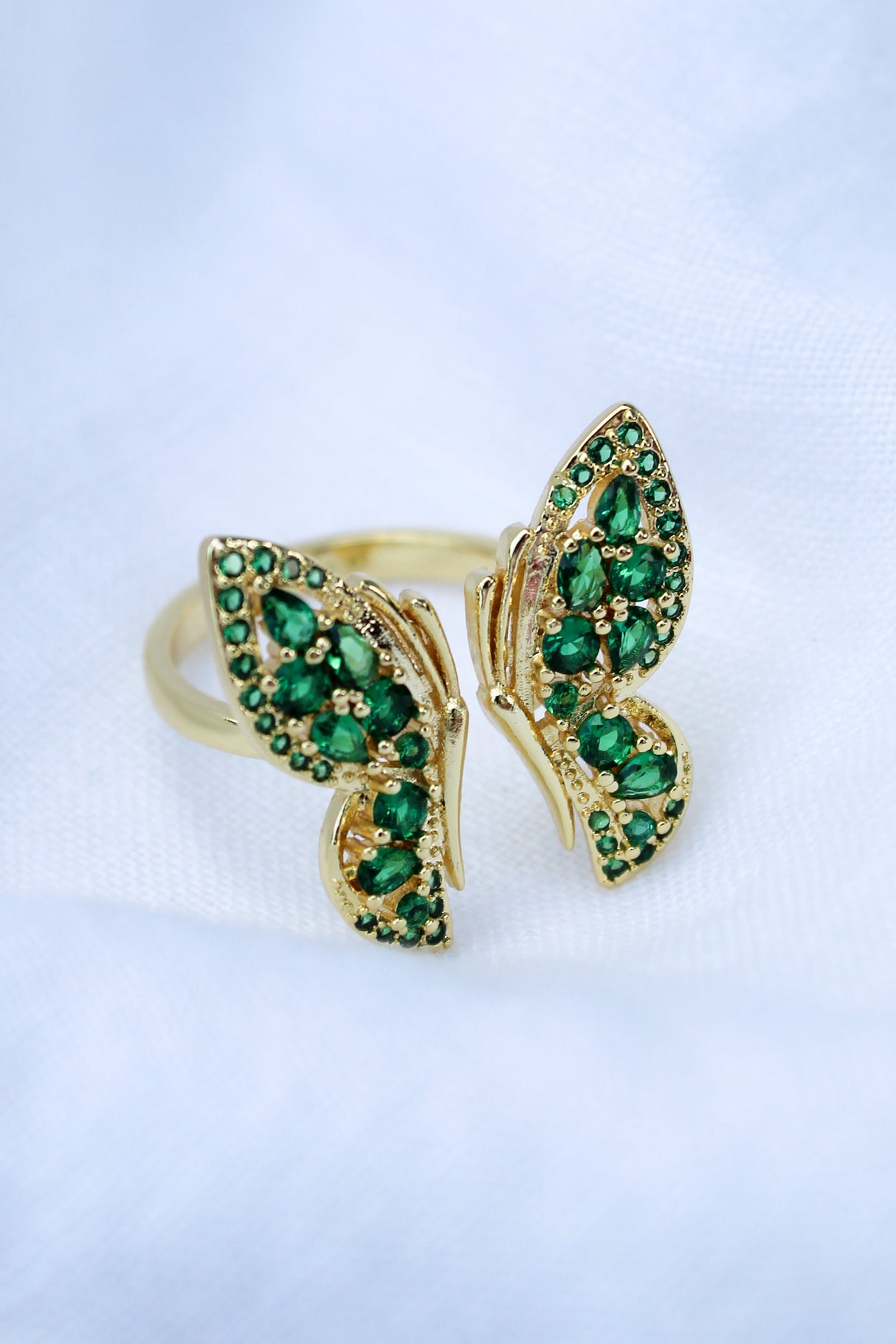 The Ezra Ring by Kinsey Designs Archives is a gold filled adjustable ring featuring two butterfly wings with small green gemstones and a .75" face, displayed on a white cloth background.