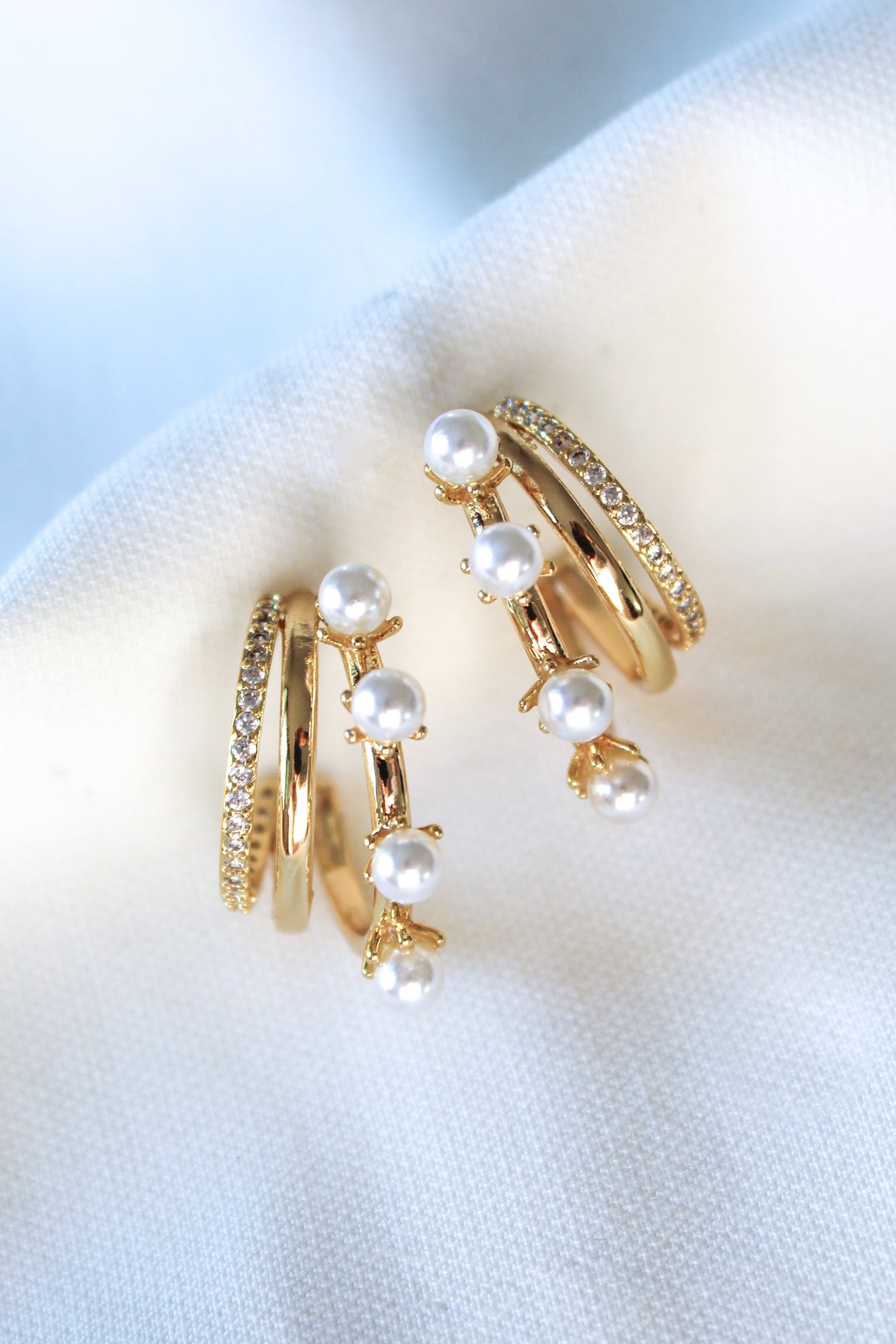 The Hattie Pearl Hoops by Kinsey Designs feature a triple-layer gold-filled design with pearl accents, adding a modern twist to classic hoops. Shown on a white fabric background.