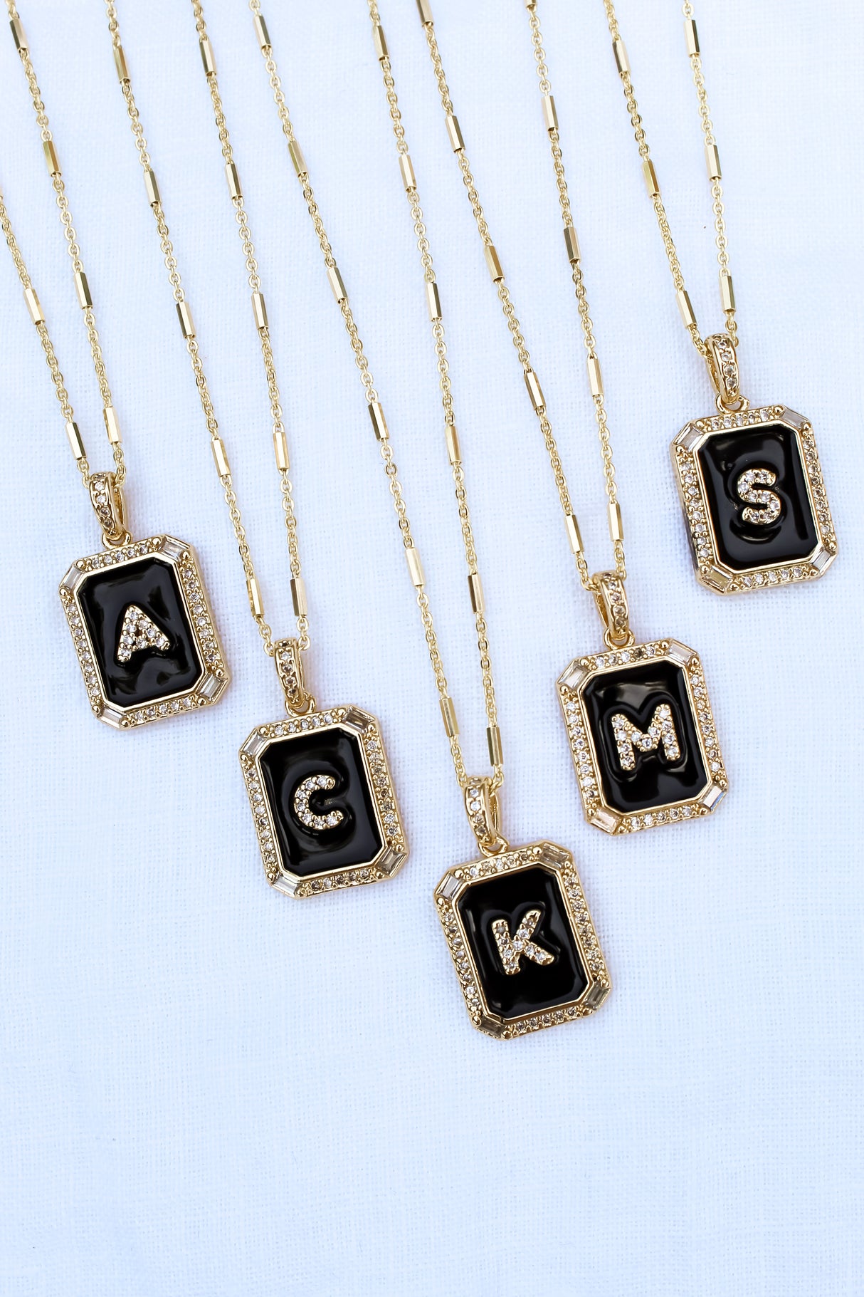 Five gold necklaces from Kinsey Designs Archives, each with a black pendant and a different silver letter—A, G, K, M, S—like the Initial Tile Necklace Ink Letter V.