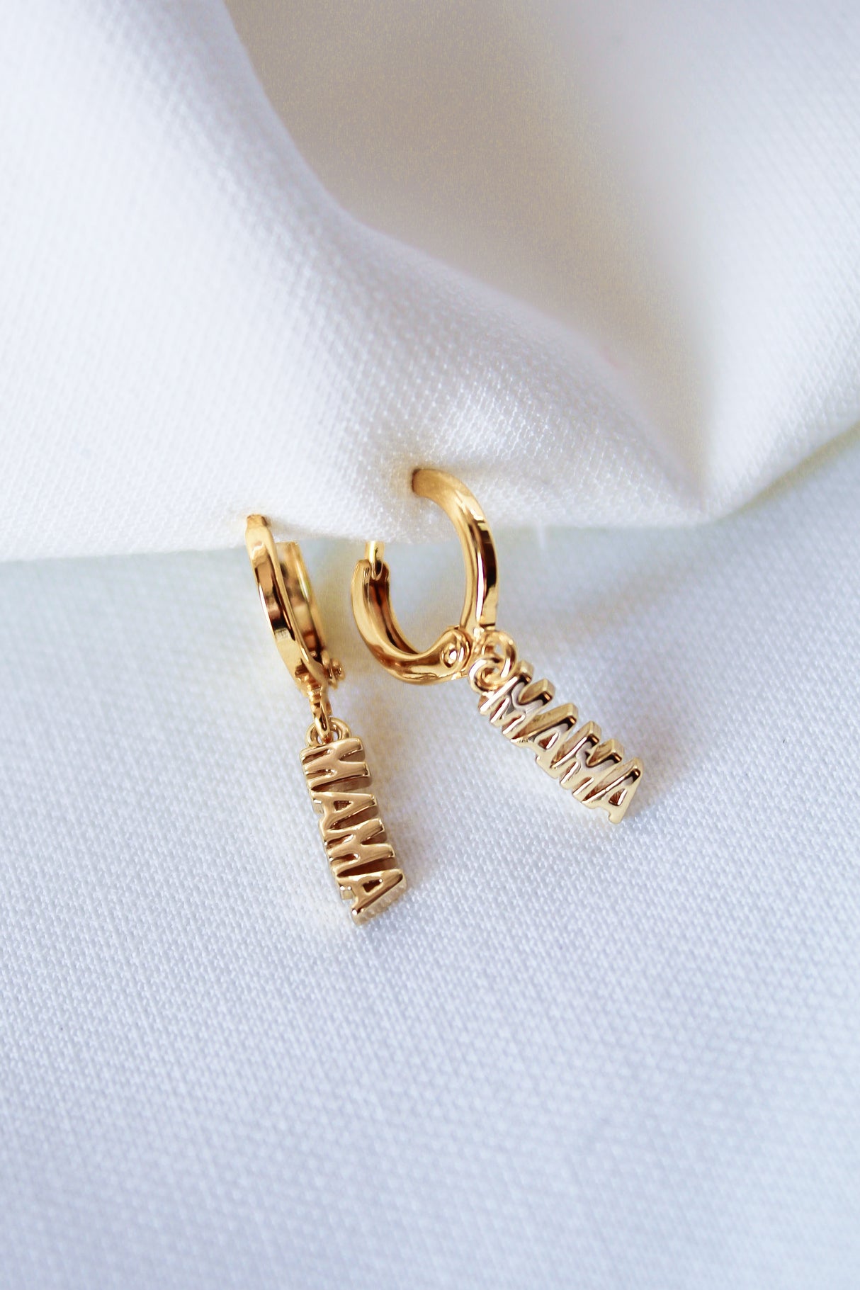 The MAMA Huggies by Kinsey Designs Archives are 1-inch gold filled earrings featuring "MAMA" letter charms, elegantly displayed on white fabric.